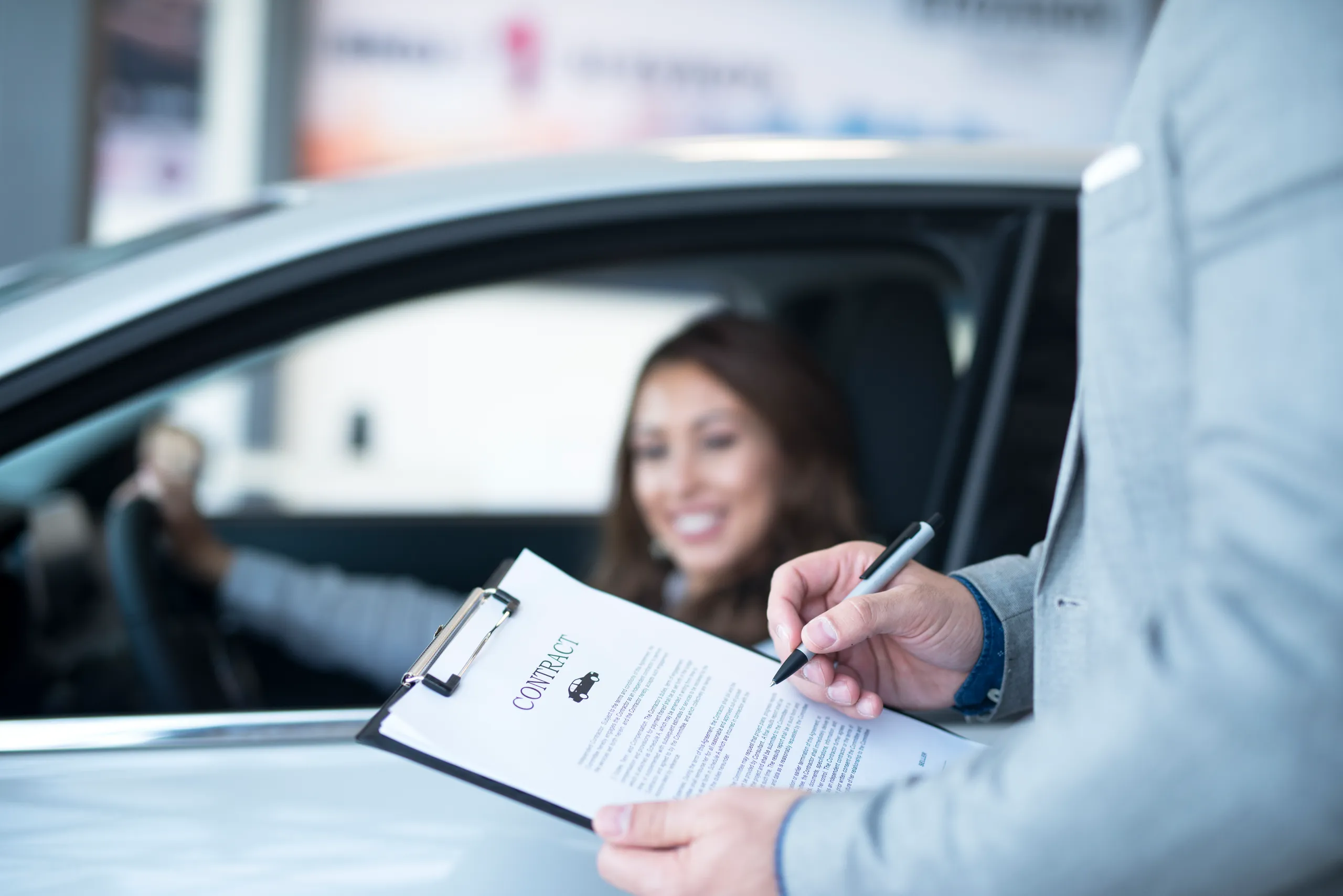 Manos firmando contrato de coche con mujer de fondo Firma de contrato de coche con mujer de fondo tras revisar el historial del coche.
